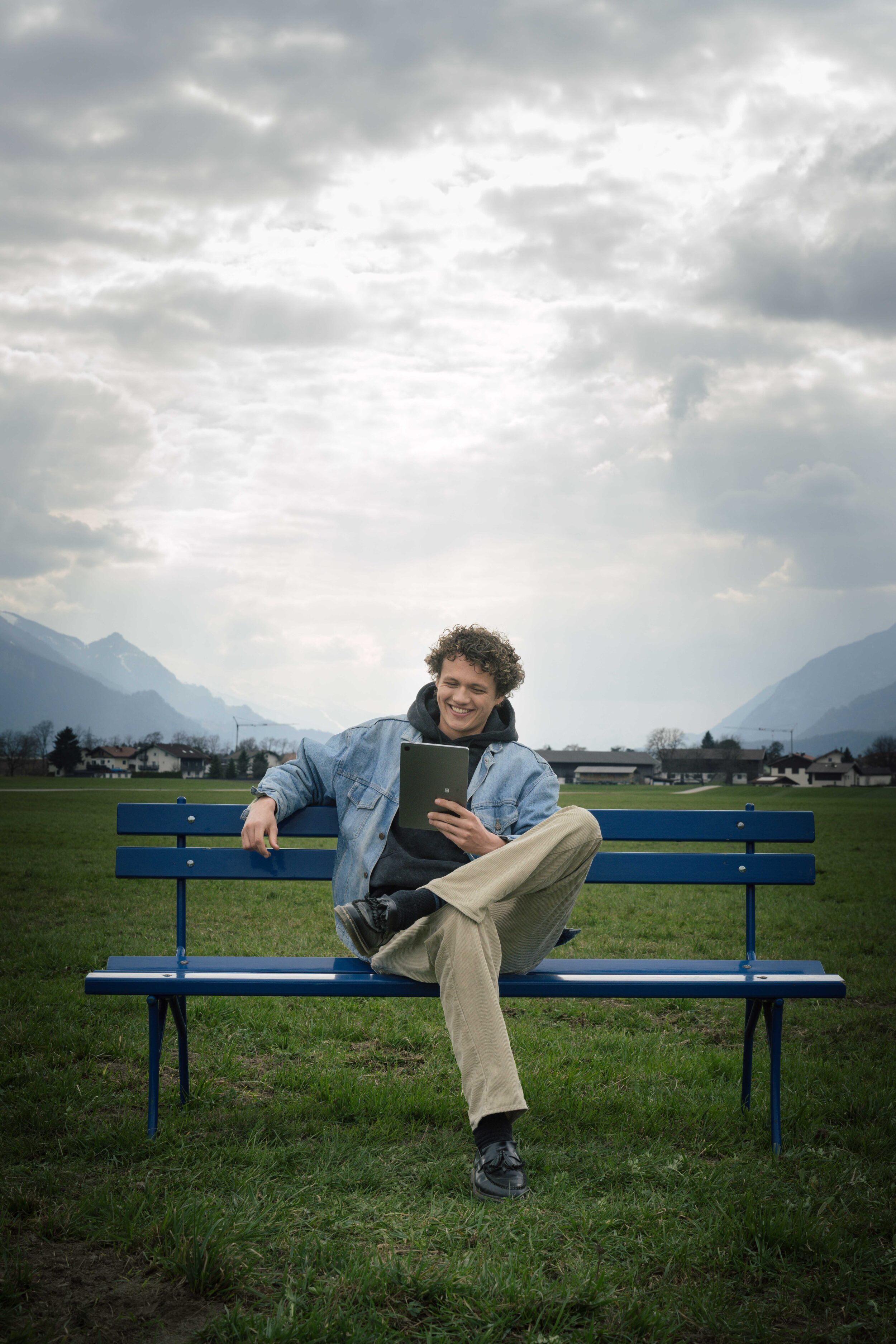 Junger Mann sitzt auf der hypoblauen Parkbank auf einem Feld und lächelt ins Tablet - freut sich über den Wohnkredit-Rechner