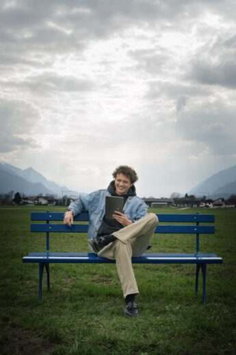Junger Mann sitzt auf der hypoblauen Parkbank auf einem Feld und lächelt ins Tablet - freut sich über den Wohnkredit-Rechner