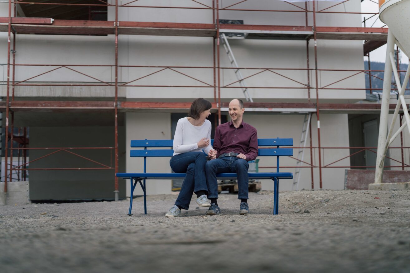 Frau und Mann sitzen lachend auf einer blauen Parkbank, die vor einem Haus mit Gerüst steht.