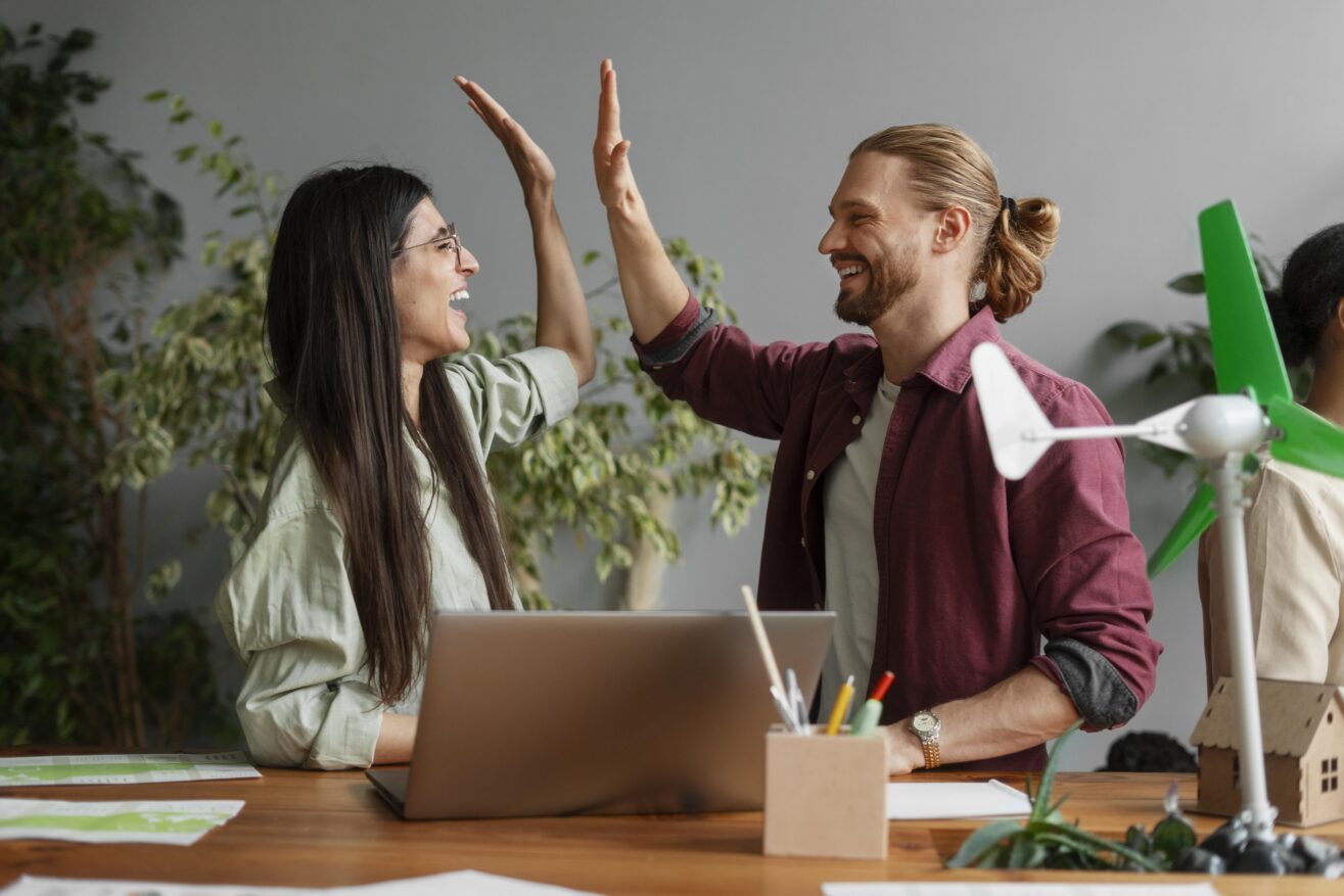 Zwei junge Menschen schauen sich gegenseitig an und klatschen in die Hände und lachen. Sie befinden sich in einem Büro, vor ihnen steht ein Laptop.
