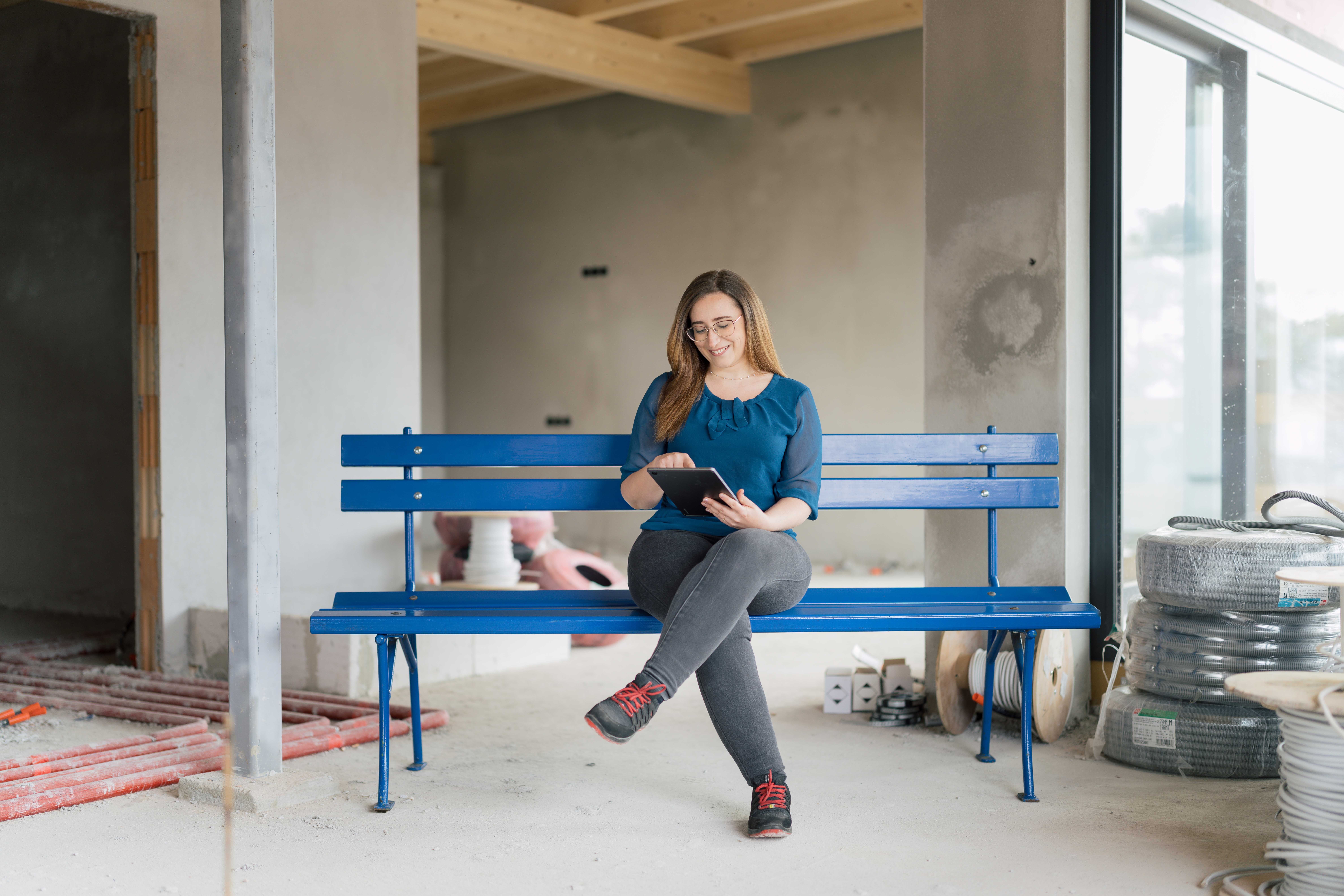 Eine Frau sitzt mit Tablett auf der hypoblauen Parkbank inmitten eines Rohbaus.