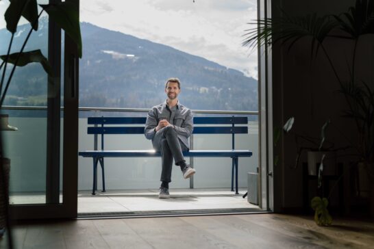 Mann sitzt auf der hypoblauen Parkbank, die auf einer Dachterrasse steht. Im Hintergrund ein Berg.