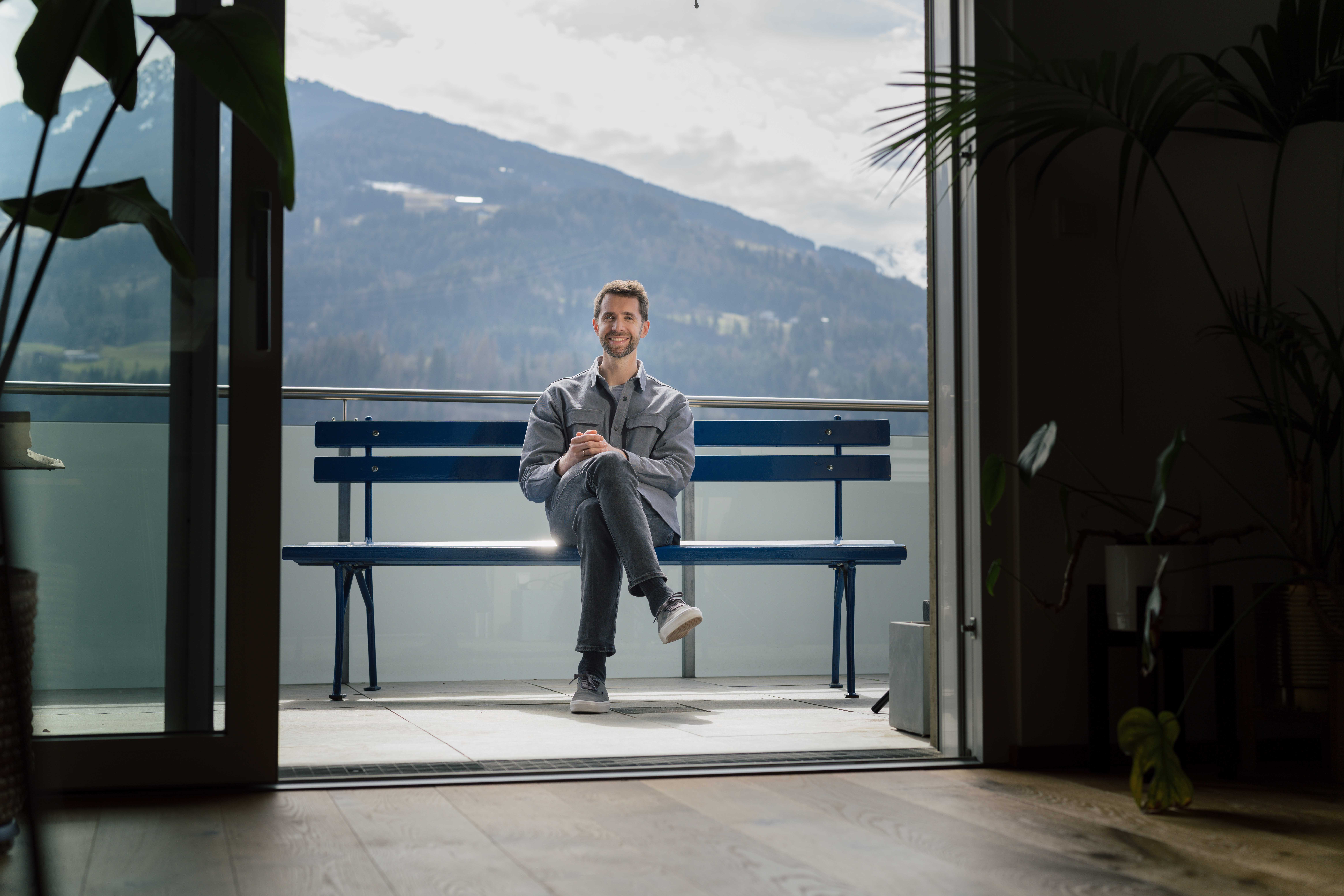 Mann sitzt auf der hypoblauen Parkbank, die auf einer Dachterrasse steht. Im Hintergrund ein Berg.